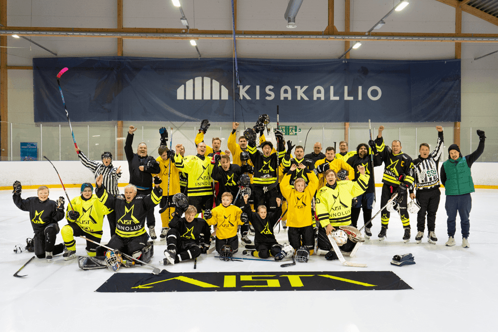Vuoden 2025 leirin yhteiskuva, jossa on suuri ryhmä aikuisia ja lapsia jääkiekkokentällä. Osa seisoo ja osa istuu tai polvistuu jäällä. Heillä on päällä kirkkaankeltaisia ja mustia pelipaitoja, joissa on Aistin keltainen tai musta tähtilogo sekä erilaisia sponsorilogoja. Osa pelaajista pitää jääkiekkomailoja kädessään, muutamilla on kypärät kädessä ja muutama istuskelee jäällä jääkiekkovarusteissaan. He poseeraavat iloisesti kameralle ja näyttävät juhlivan, moni pitää käsiä tai mailaa ylhäällä. Etualalla on iso musta lippu, jossa on keltainen tähti ja teksti 