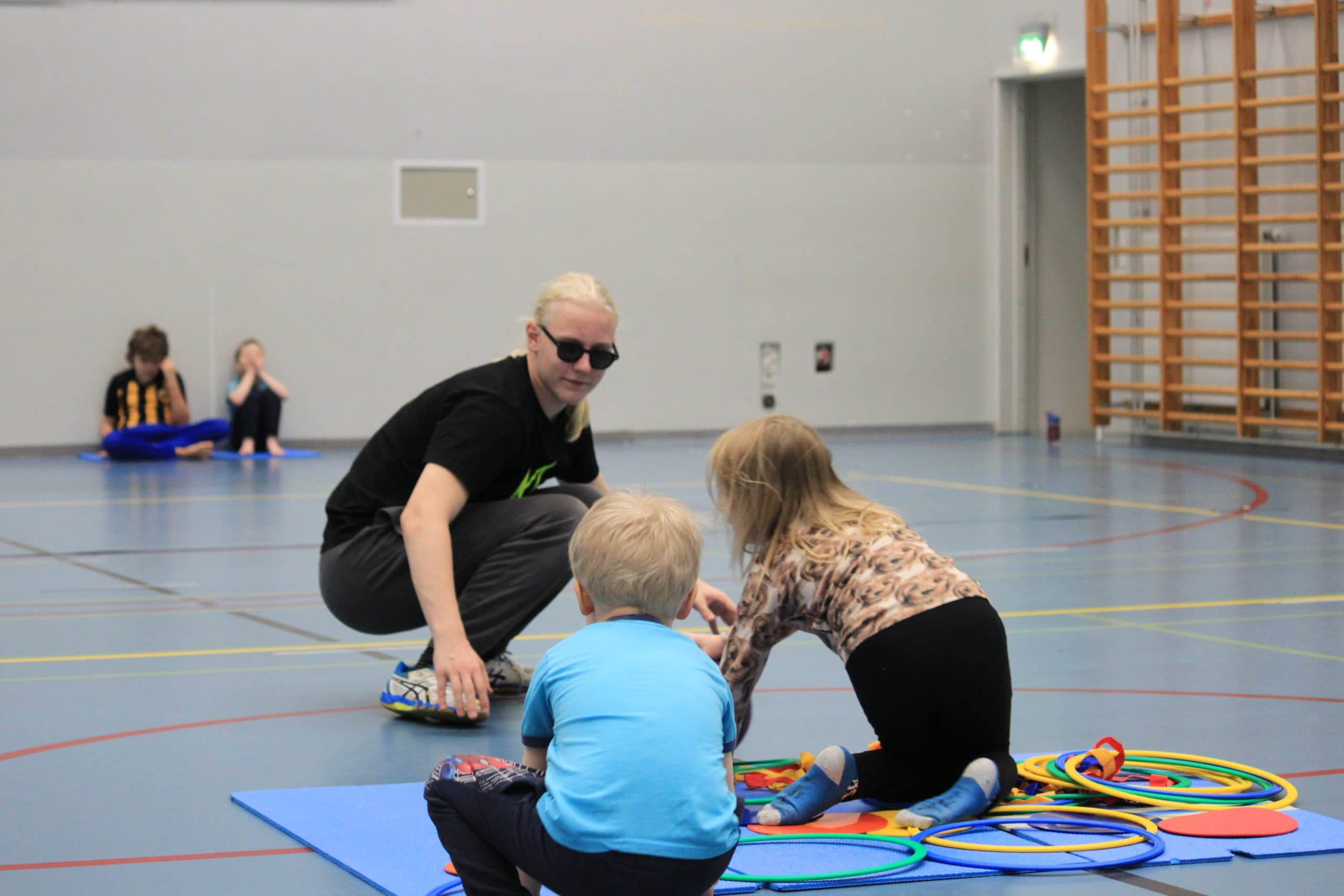 A young male instructor crouches to guide two small children in their play. The children play on a blue exercise mat scattered with colourful toy rings.