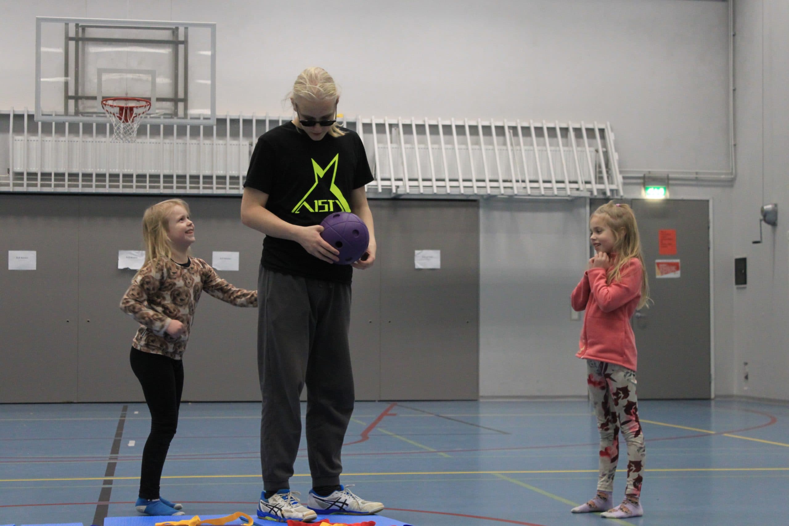 A young male instructor stands holding a children's goalball, guiding two girls. The girls stand on either side of the instructor and smile happily.