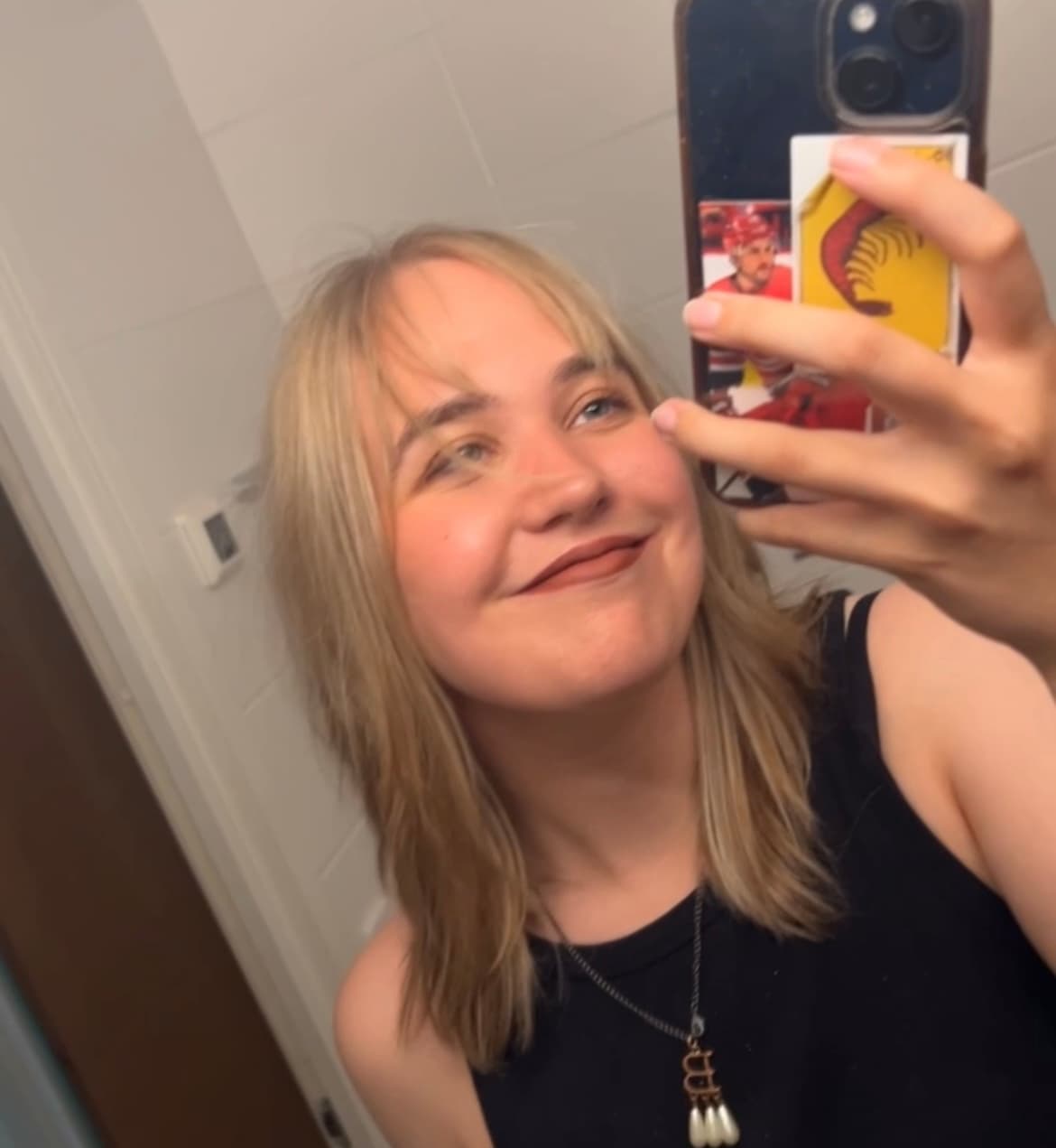 A relaxed photo where a young woman takes a mirror selfie. She has fair, shoulder-length hair and smiles broadly. She wears a black sleeveless top and a necklace, and holds a phone whose back cover has a trading card of hockey player Nino Niederreiter.