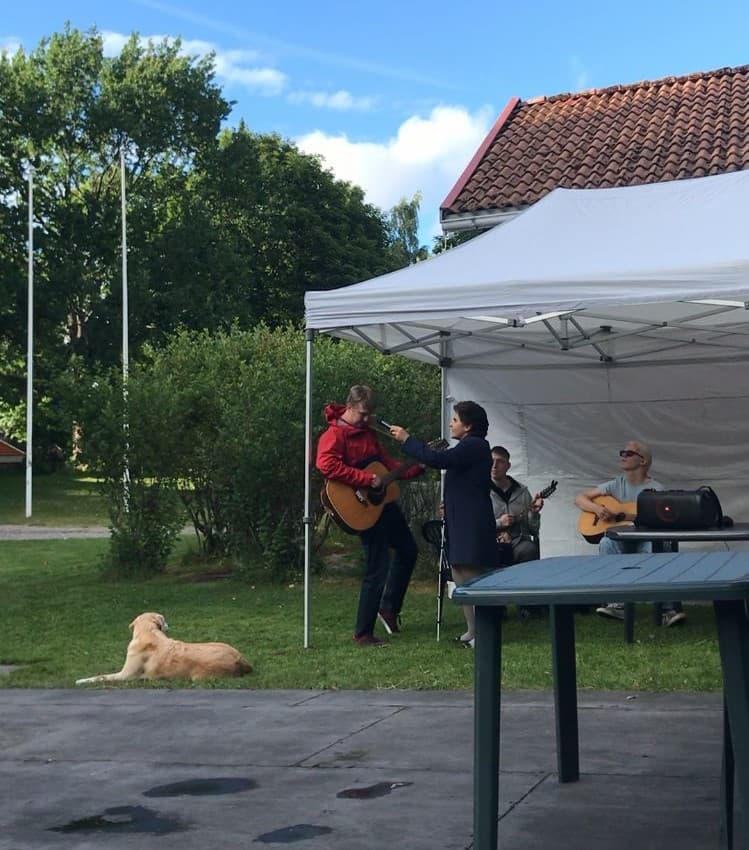 Poikien bändi (Eemi, Akseli ja Samuli) teltassa, Azra mikinpitelijänä, bändäri-Helmi ja yksi kuvaajan ja teltan väliin jäänyt tyhjä pöytä.
