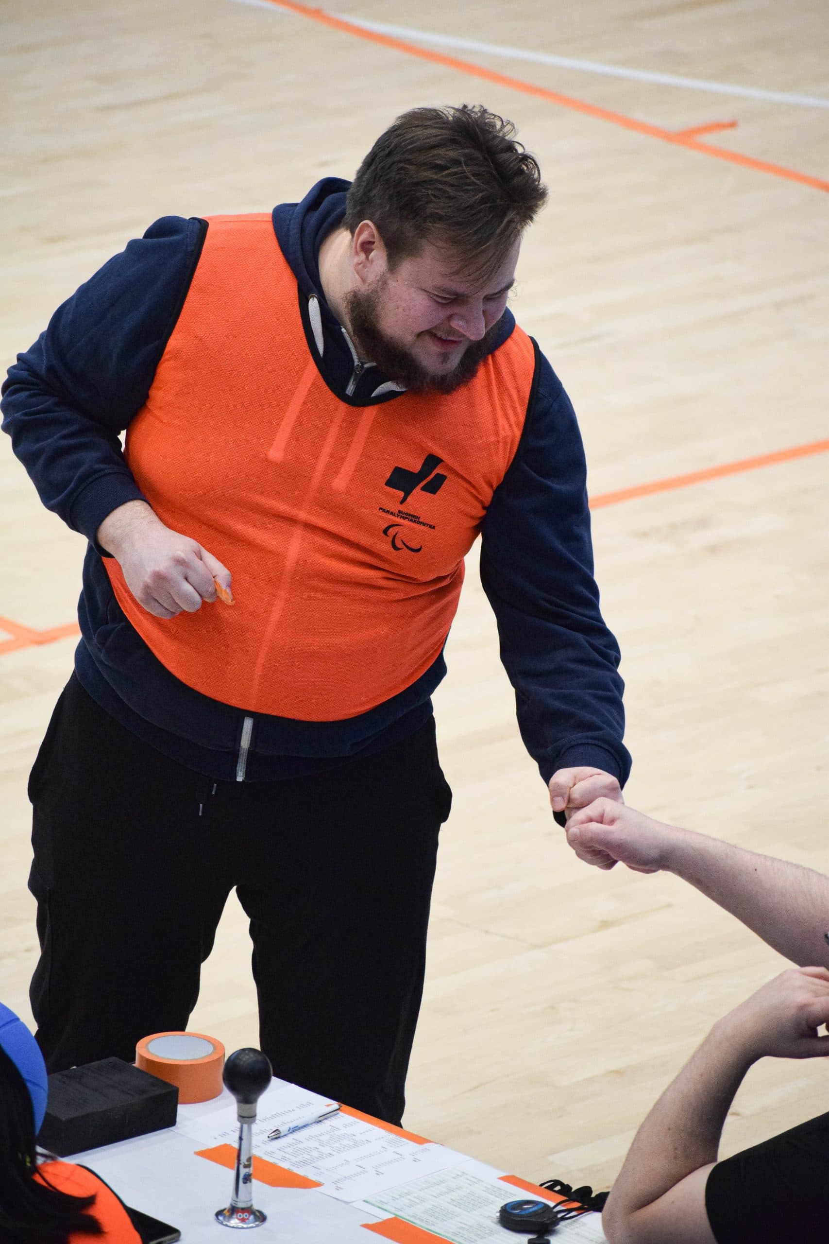 Kuvassa virnistävä Pete lyömässä kiviä (nyrkkiä) yhteen oranssissa vapaaehtoisliivissä kuvan ulkopuolelle rajautuvan tuomaripöydän käden kanssa.