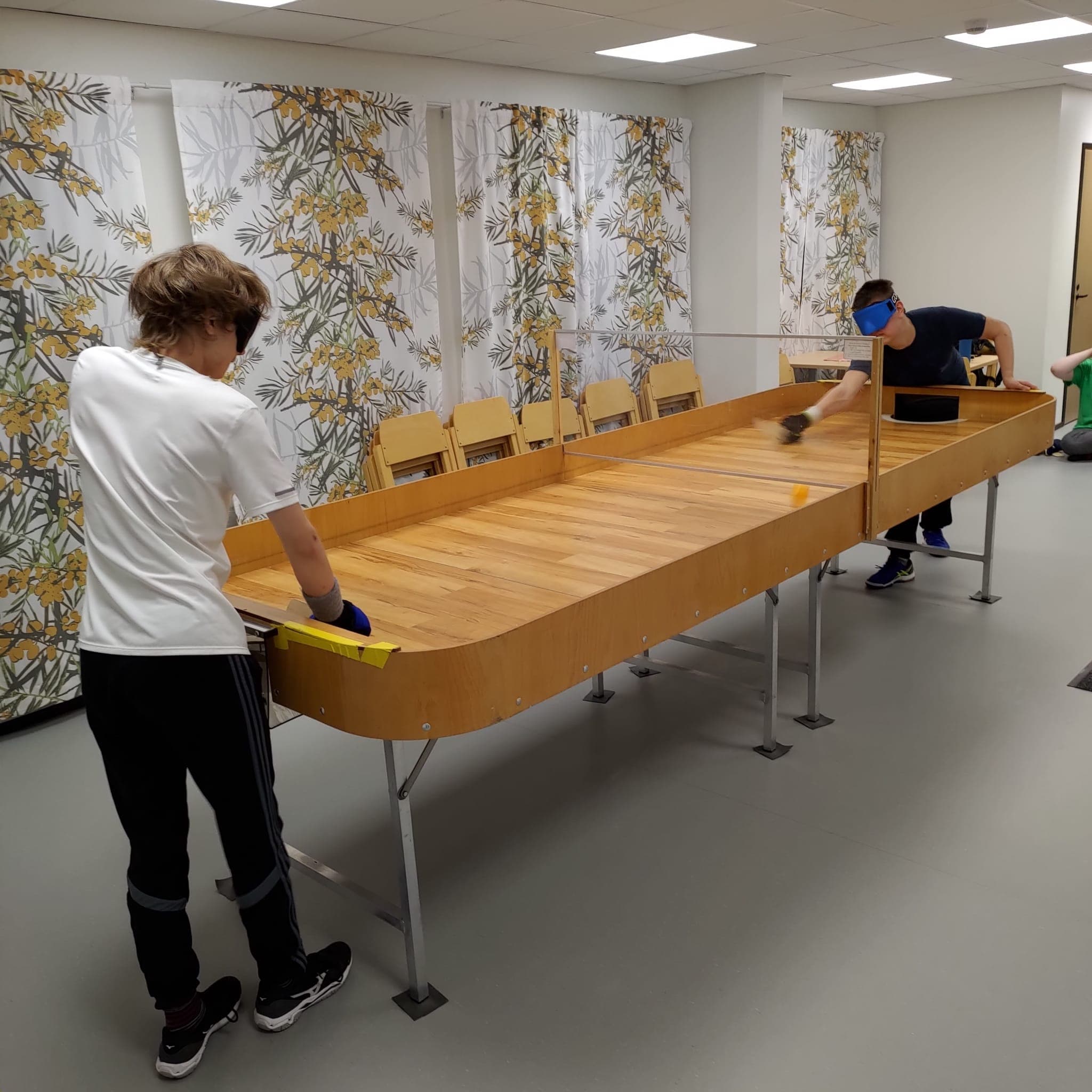 Pelipöydän kulmalta otettu kuva, jossa kaksi pelaajaa pelipöydän molemmissa päissä pelilaseineen ja puisine mailoineen.Takimmainen pelaaja on juuri lyönyt palloa. 