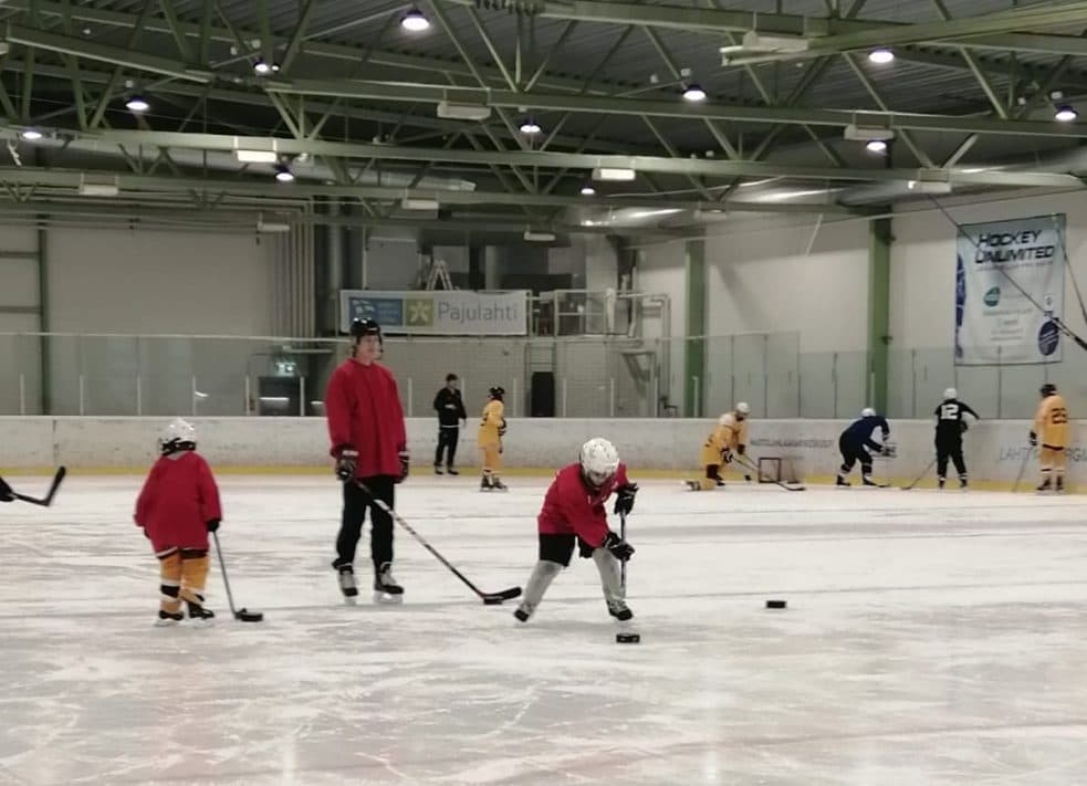 Pienikokoinen lapsi luistelee kiekon kanssa. Taustalla valmentaja seuraa ja toinen lapsi luistelee kiekon kanssa.