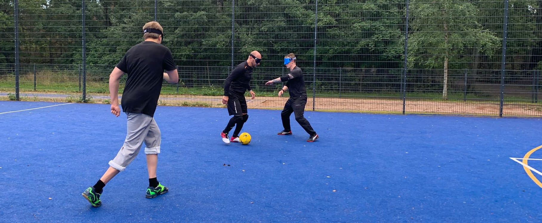 Pelitilannekuva näkövammaisjalkapallosta. Pelaaja etenee pallon kanssa. Puolustaja on käden etäisyyden päässä vastassa ja toinen pelaaja juoksee etualalla rinnalla.