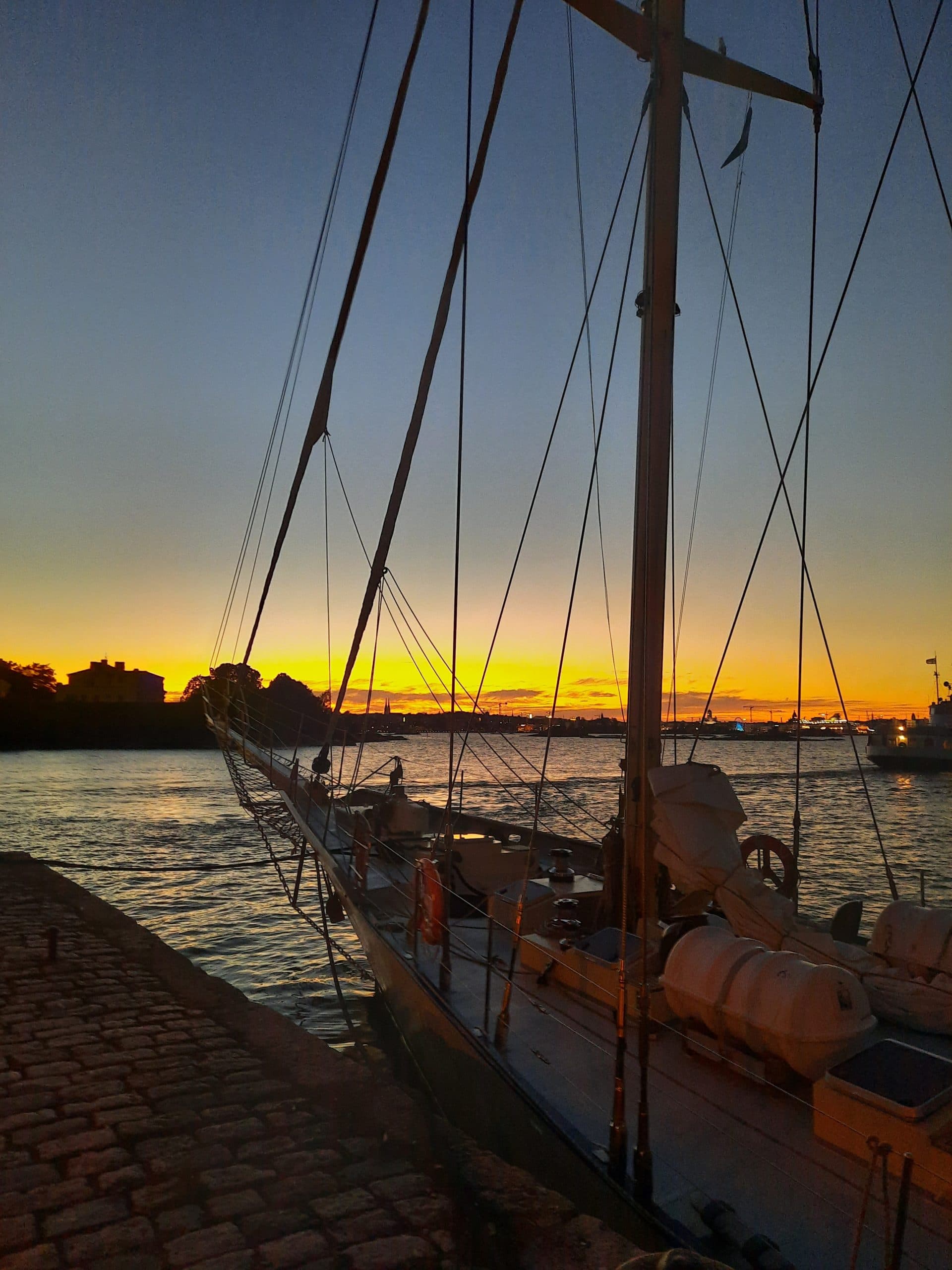 Purjelaiva Kuunari Helena satamassa. Kuvassa näkyy laivan etu puolisko ja etumasto. Taustalla auringonlasku ja hämärtyvä ilta lahdenpoukamassa.