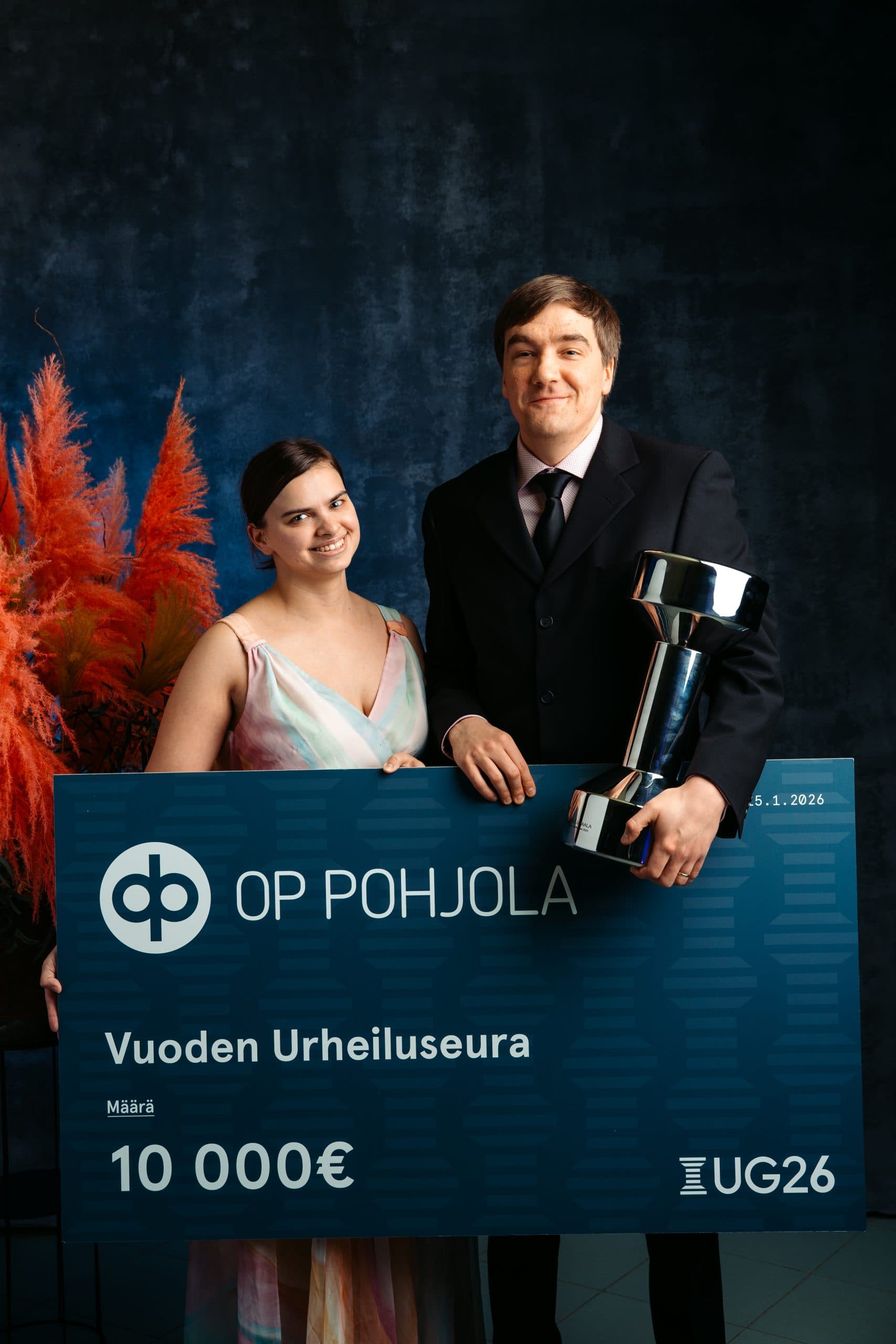 Markus Tihumäki and Anniina Tihumäki, festive and smiling, holding a giant prize cheque in front of them, with Markus also cradling the 'Poika' — the Uuno statue — under his arm.