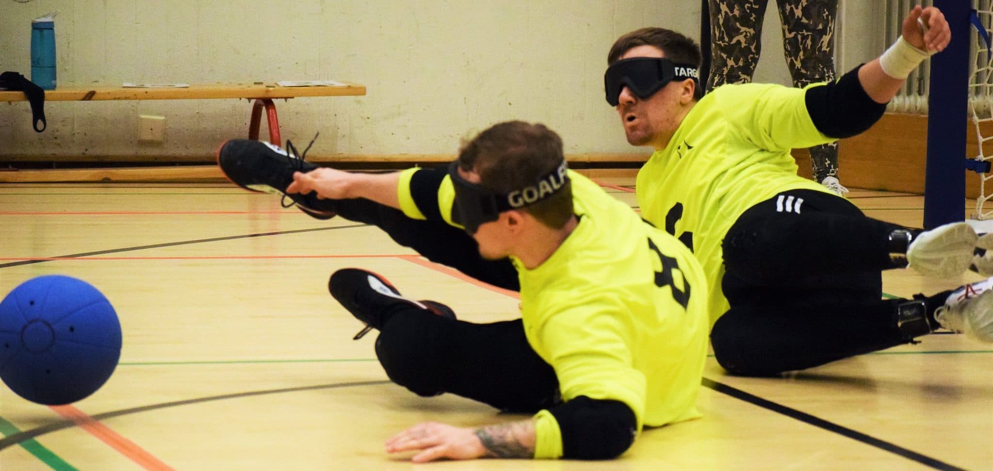 An action shot from a goalball game where the centre dives to block the approaching ball. The wing is visible in the background backing up the block.