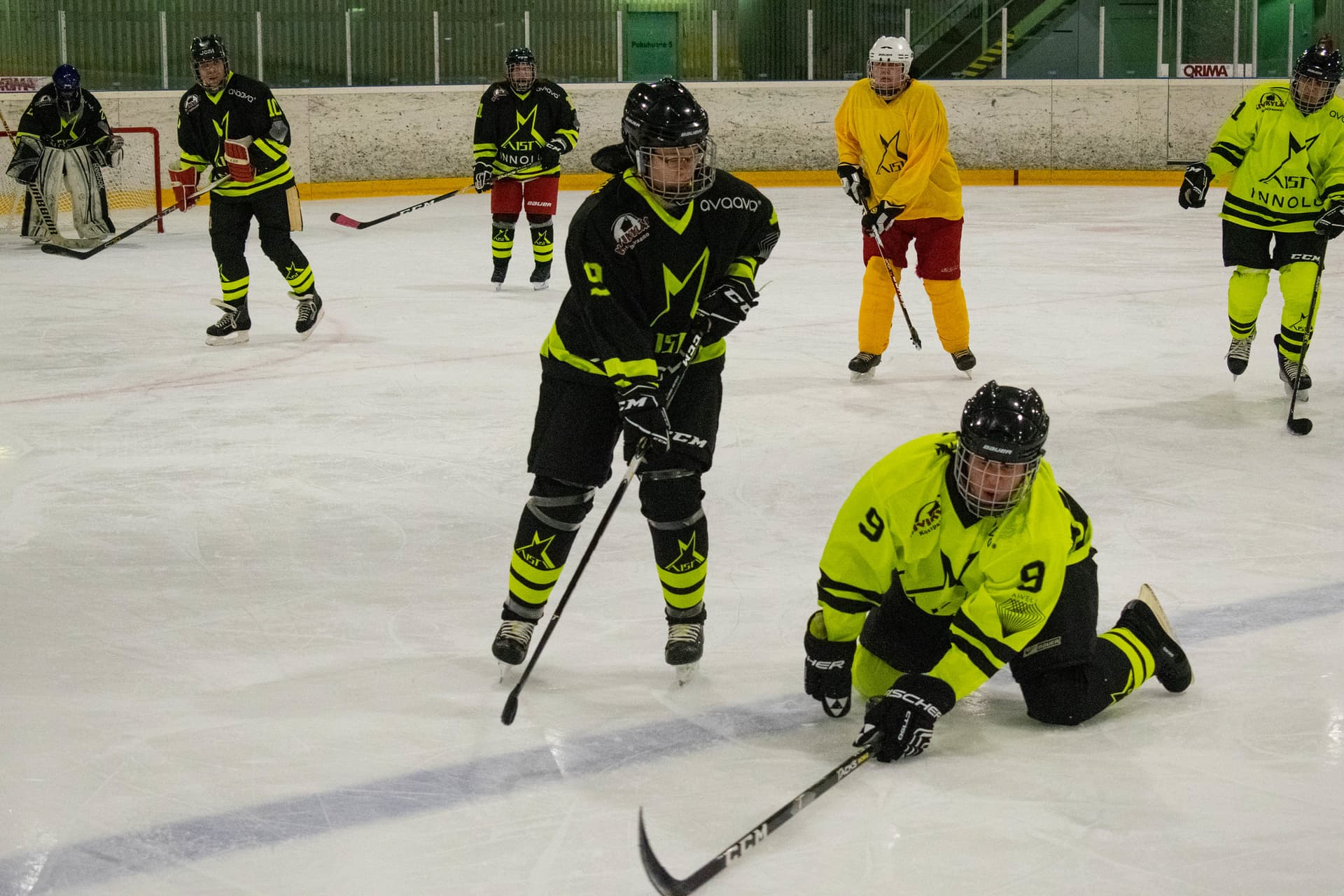 Tilannekuva näkövammaiskiekko-ottelusta jossa fluminopaitainen pelaaja numero 9 liukuu kohti kameraa polvillaan oletettavasti kiekon perässä ja mustapaitainen pelaaja luistelee vieressä. Taustalla näkyy mustapaitainen maalivahti ja kaksi puolustajaa.