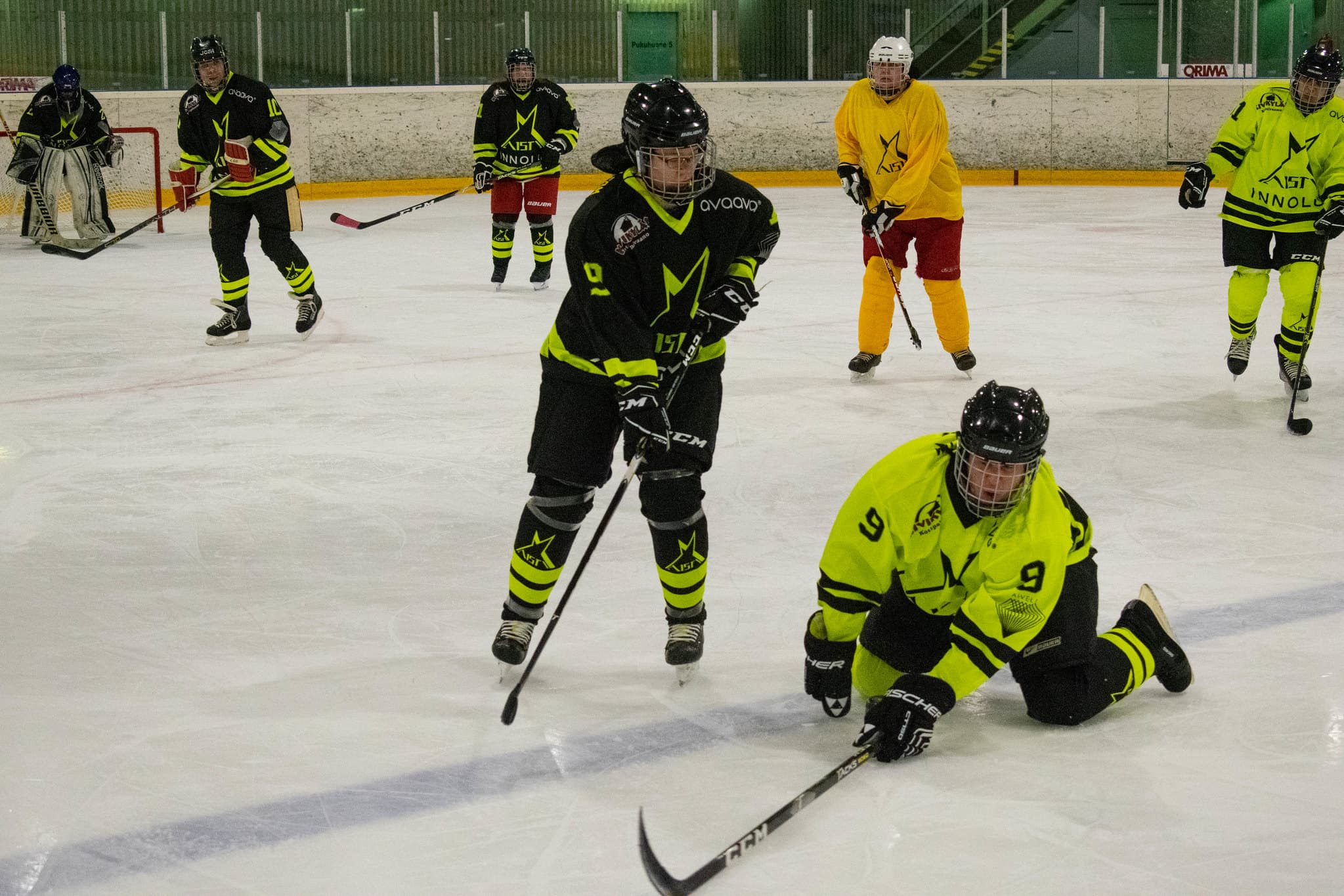 Tilannekuva näkövammaiskiekko-ottelusta jossa fluminopaitainen pelaaja numero 9 liukuu kohti kameraa polvillaan oletettavasti kiekon perässä ja mustapaitainen pelaaja luistelee vieressä. Taustalla näkyy mustapaitainen maalivahti ja kaksi puolustajaa.