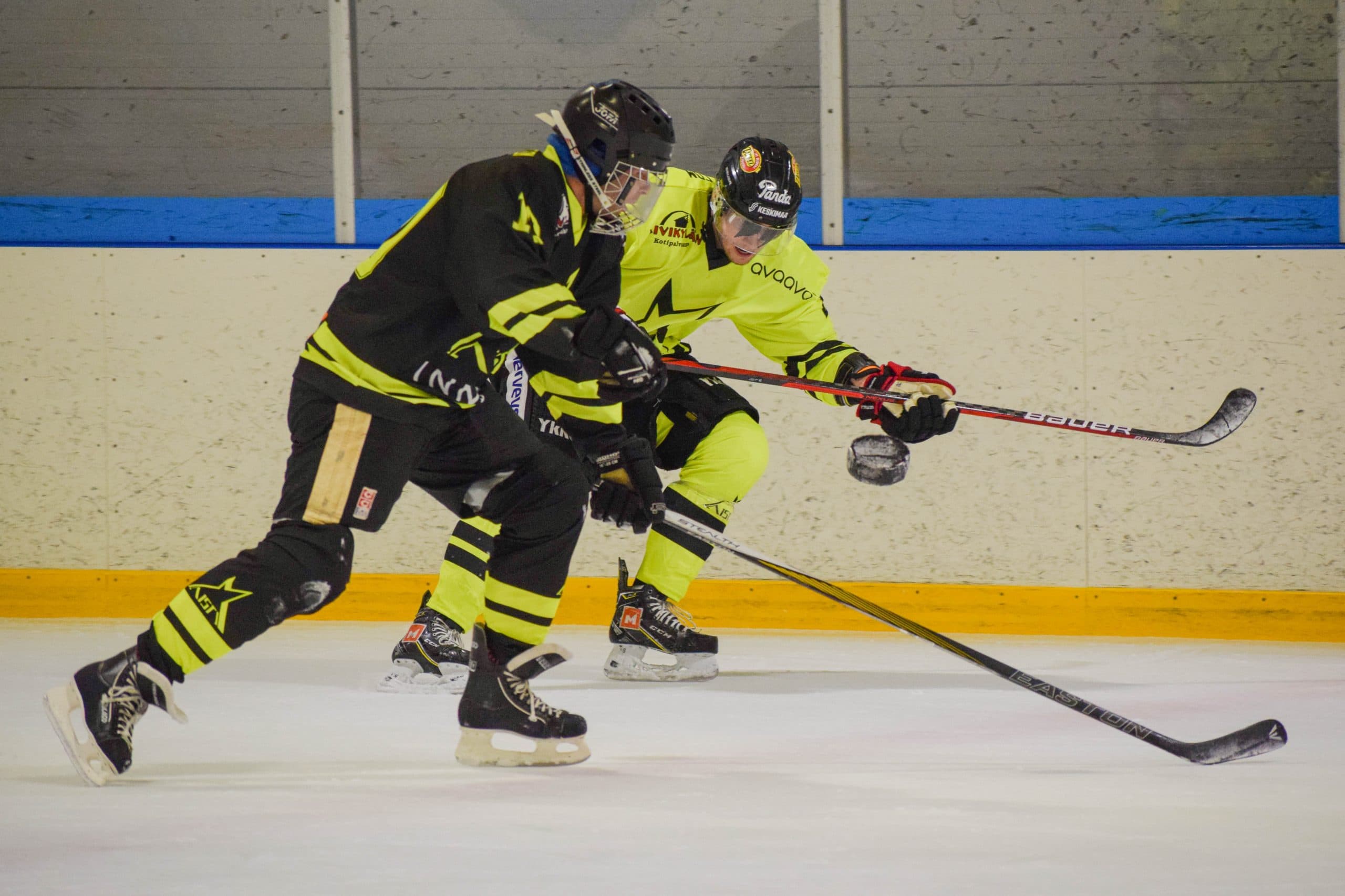 An action shot from a blind ice hockey match where Aisti Sport players in flumino and black jerseys skate at speed along the boards chasing the puck. Both players have their sticks leaning forward and the black puck is in the air in front of them.