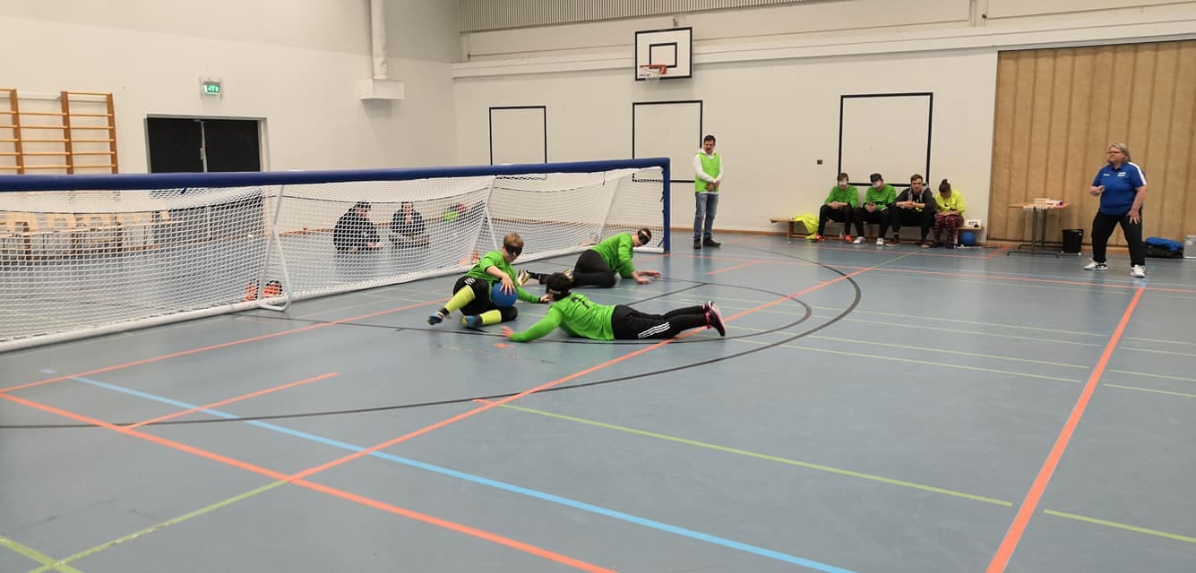 Pelitilannekuva, jossa Aisti Openin laitapelaaja syöksyy torjuntaan sentterin taakse. Taustalla näkyy vaihtopenkki, jossa valmentaja ja vaihtopelaajat seuraavat tilannetta.