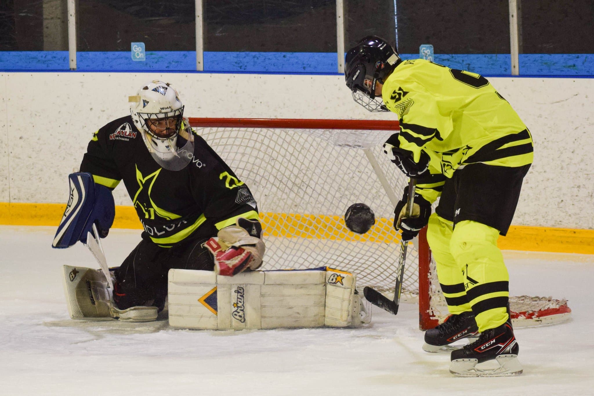 The black team's goalkeeper, eyes fixed on the puck flying in the air in front of the goal. A player from the yellow team lifts the puck with a backhand towards the top corner of the goal.