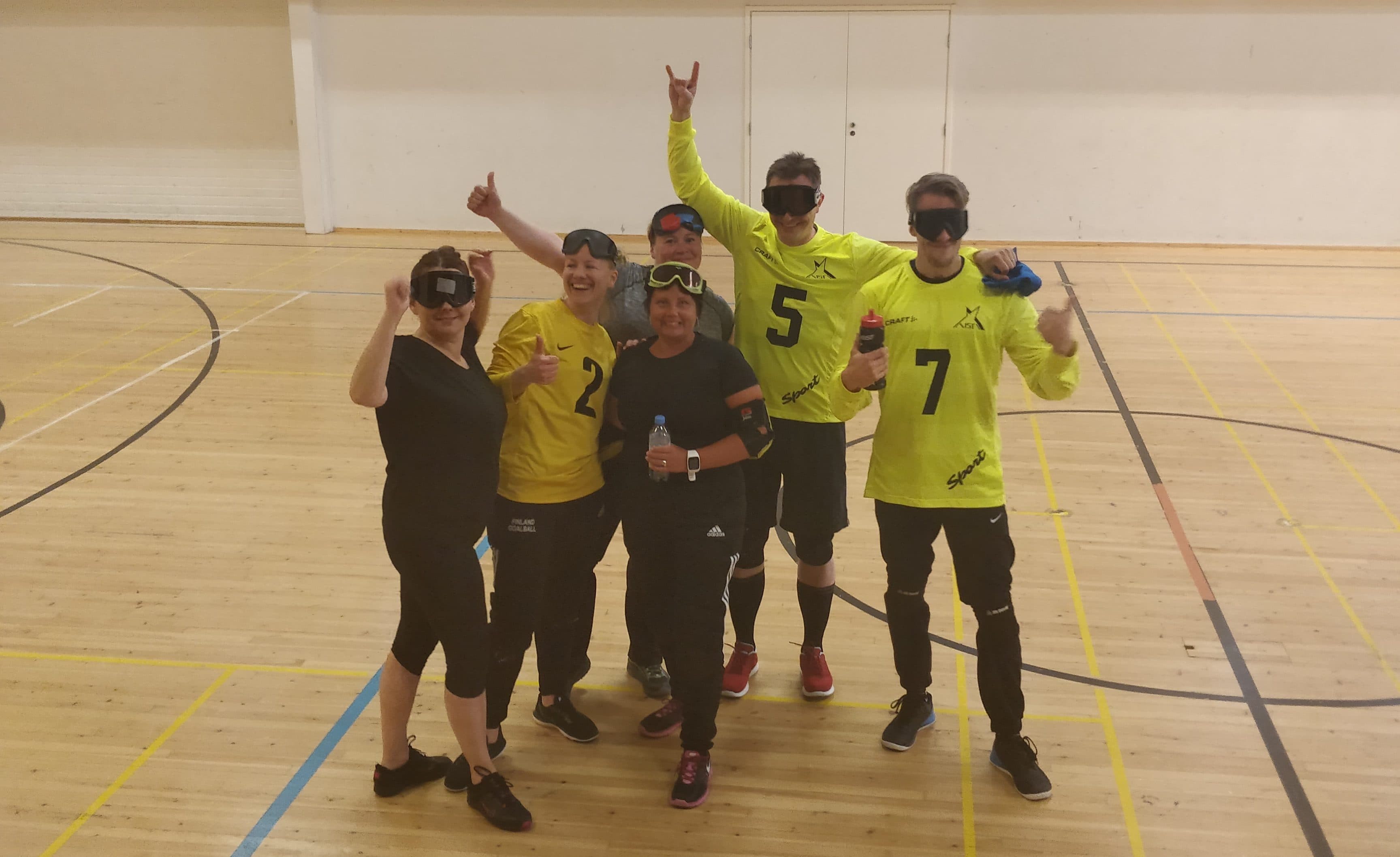 A photo where goalball newcomers laugh on the court in playing gear. Some give a thumbs-up, some have goalball eyeshades over their eyes, some on their foreheads.