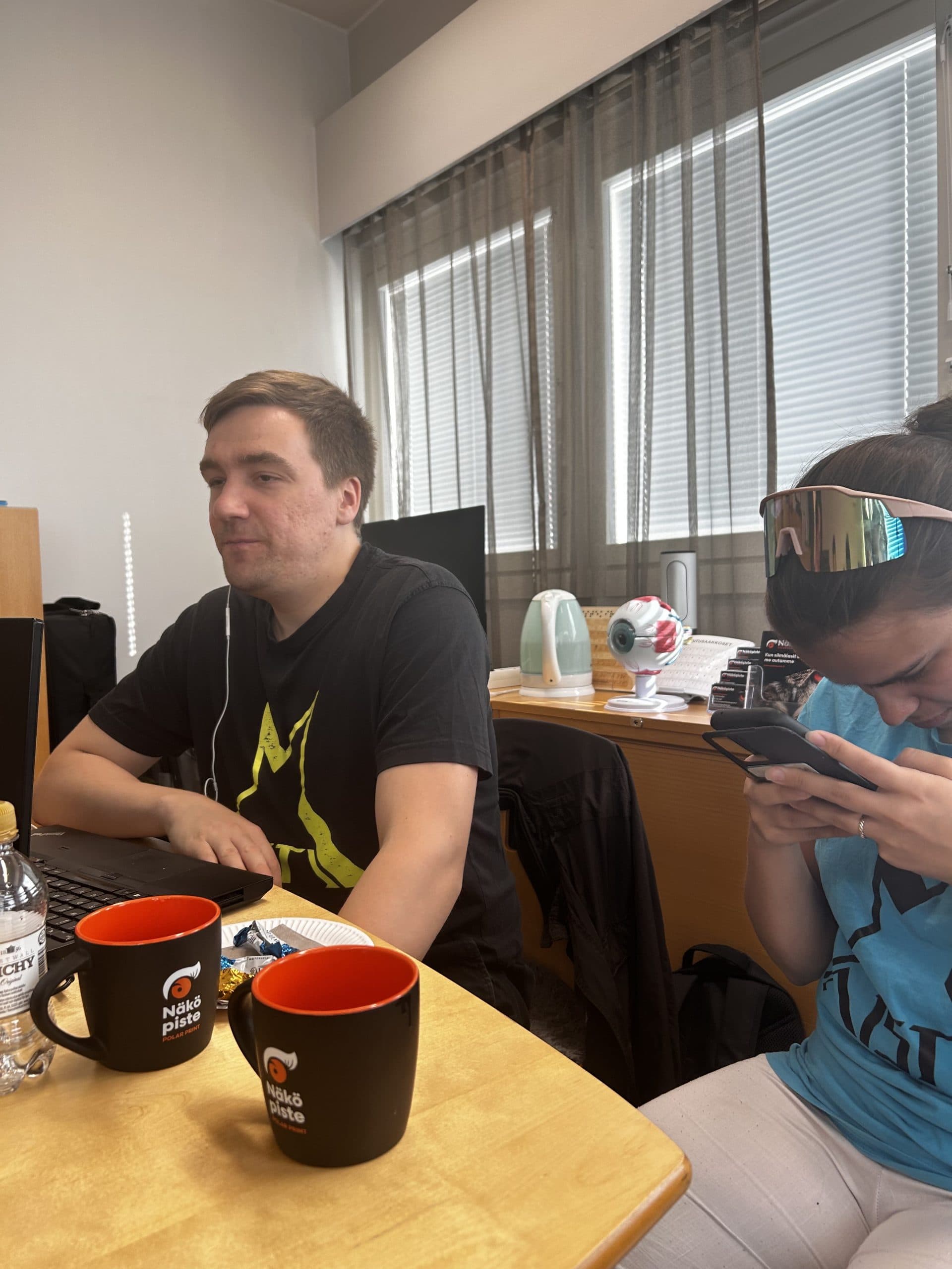 Markus ja Ansku istuvat Aisti Sportin paidat päällään pöydän ääressä Polar Printin logolla varustetut kahvikupit edessään ja Markuksella läppäri pöydällä edessään.