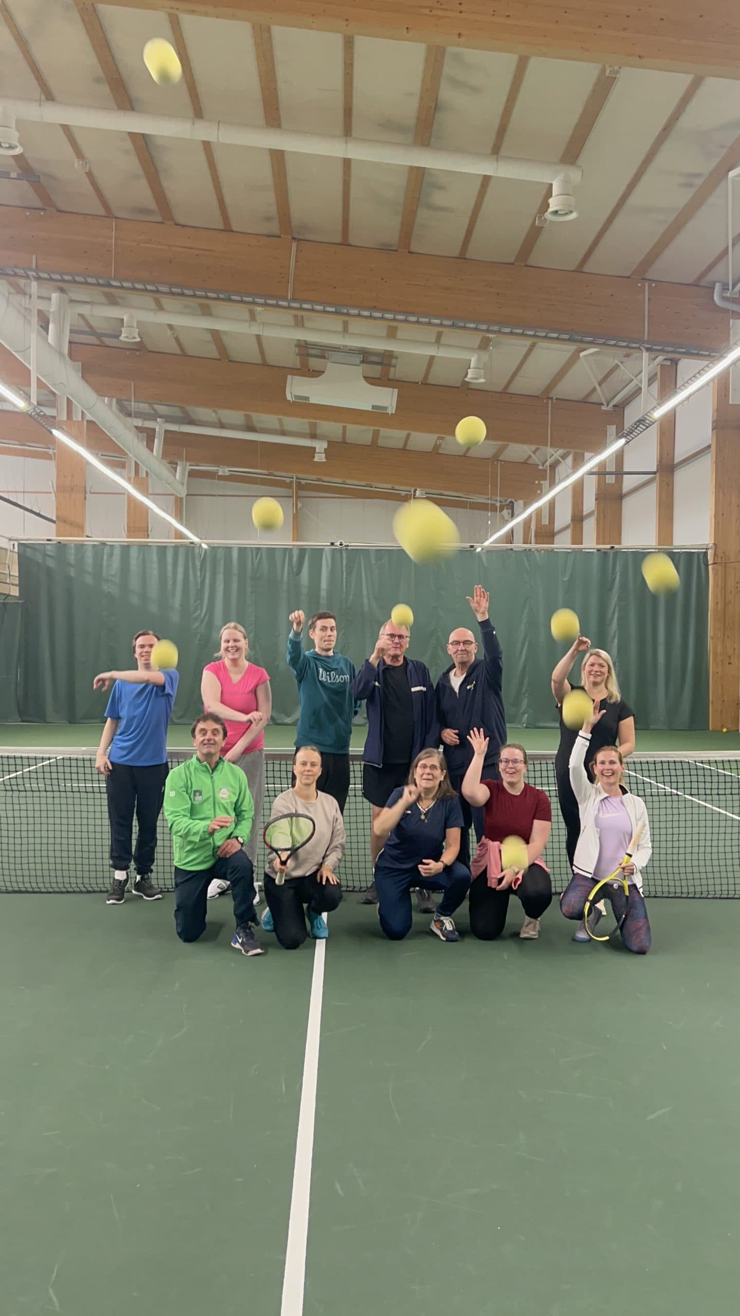 People on a tennis court in front of the net in two rows. Some players hold tennis rackets, and everyone throws tennis balls towards the camera. Approaching tennis balls are visible all over the image.