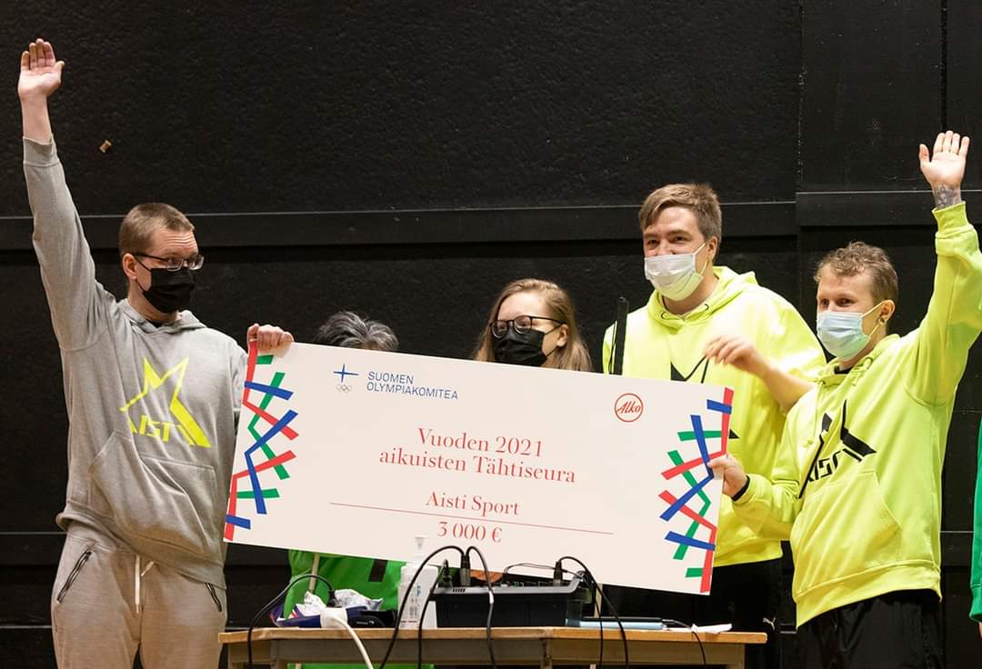 A photo taken from the stage, where the Aisti board celebrates with arms raised, holding a large cardboard prize cheque.