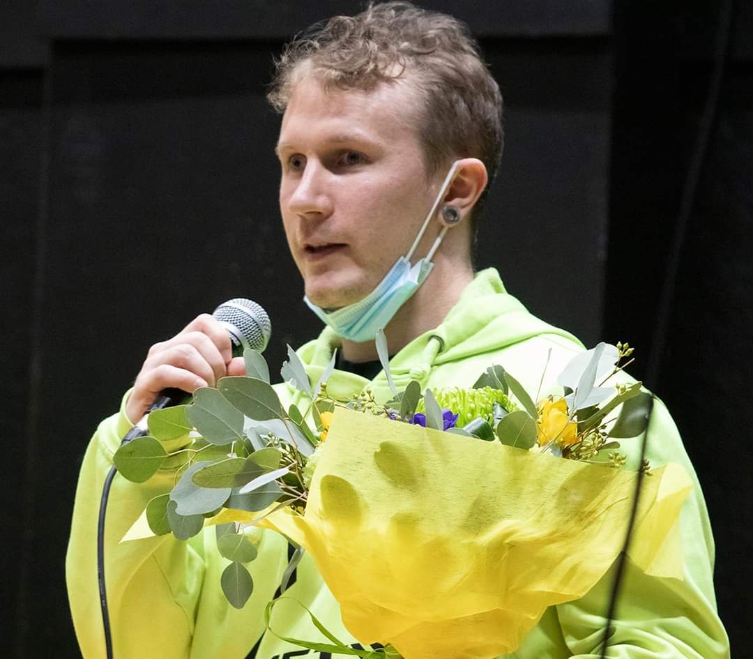 Puheenjohtaja Miika Honkanen puhuu mikrofoniin Aistin flumino huppari yllään ja kukkakimppu vasemmassa kädessään.
