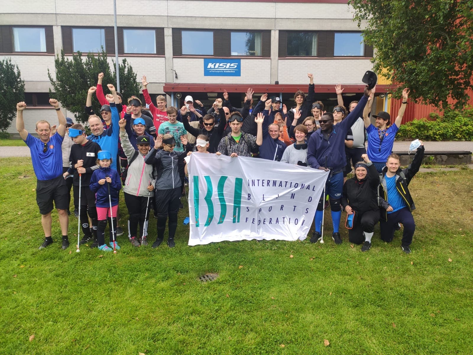 Näkövammaisjalkapalloleirin ryhmäkuva, jossa hymyileviä lapsia ja aikuisia ulkona otetussa valokuvassa.