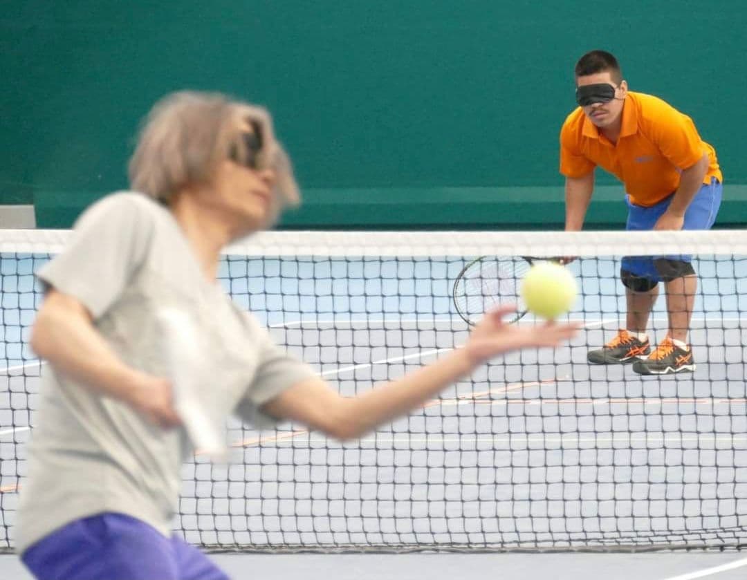 An action shot from a blind tennis match. A man in a light jersey is just tossing the ball up to serve, and opposite, on the other side of the net, a man in an orange jersey is ready to return. Both players wear black eyeshades over their eyes.