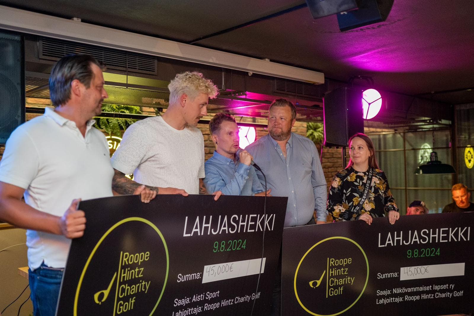 Viisi ihmistä, jotka seisovat lavalla ja pitävät suuria lahjashekkejä käsissään. Shekeissä lukee "LAHJASHEKKI" ja päivämäärä 9.8.2024. Shekkien summa on 45 000 euroa. Ensimmäisessä shekissä saajana on "Aisti Sport" ja toisessa "Näkövammaiset lapset ry". Molemmissa lahjoittajana on "Roope Hintz Charity Golf". Taustalla näkyy ravintolaympäristö ja värikkäitä valoja.