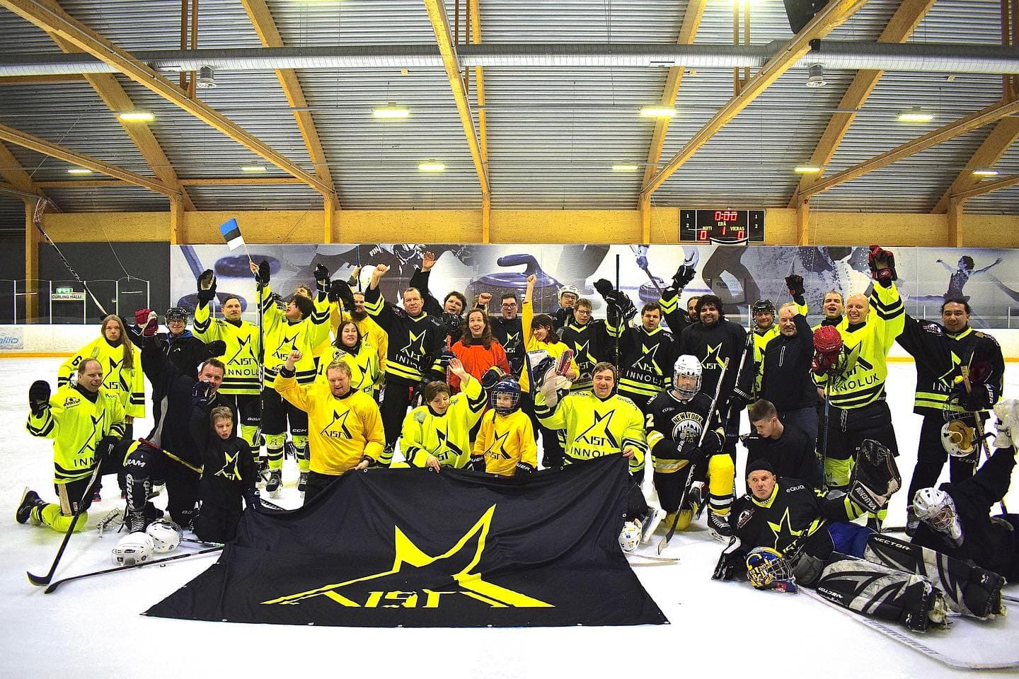 The participants of the 2023 camp posing in a group photo in an ice hockey rink. People are dressed in bright yellow and black jerseys bearing the large Aisti star logo; some jerseys also carry sponsor logos. Some players kneel or sit on the ice, wearing hockey gear such as helmets and sticks. The participants are of various ages. In the background, the rink boards with large pictures of ice hockey players. In front of the team is a large black flag with a yellow star and text.