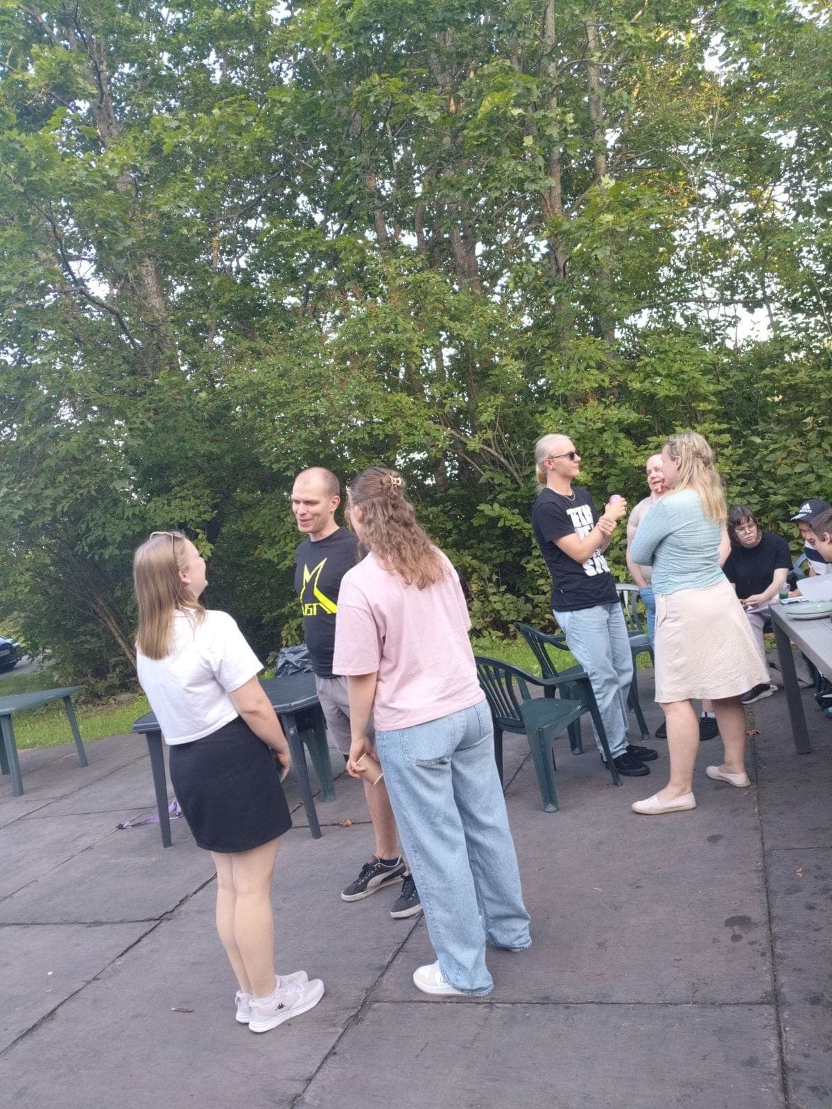 Kuva juhlivasta Aistin jäsenistöstä ulkona terassilla. Etualalla kaksi nuorta naista ja Aistin t-paitaan sonnustautunut mies juttelevat ja taustalla näkyy kaksi muuta kolmen hengen ryhmää juttelemassa. Taustalla lehtipuita terassialueen reunassa.