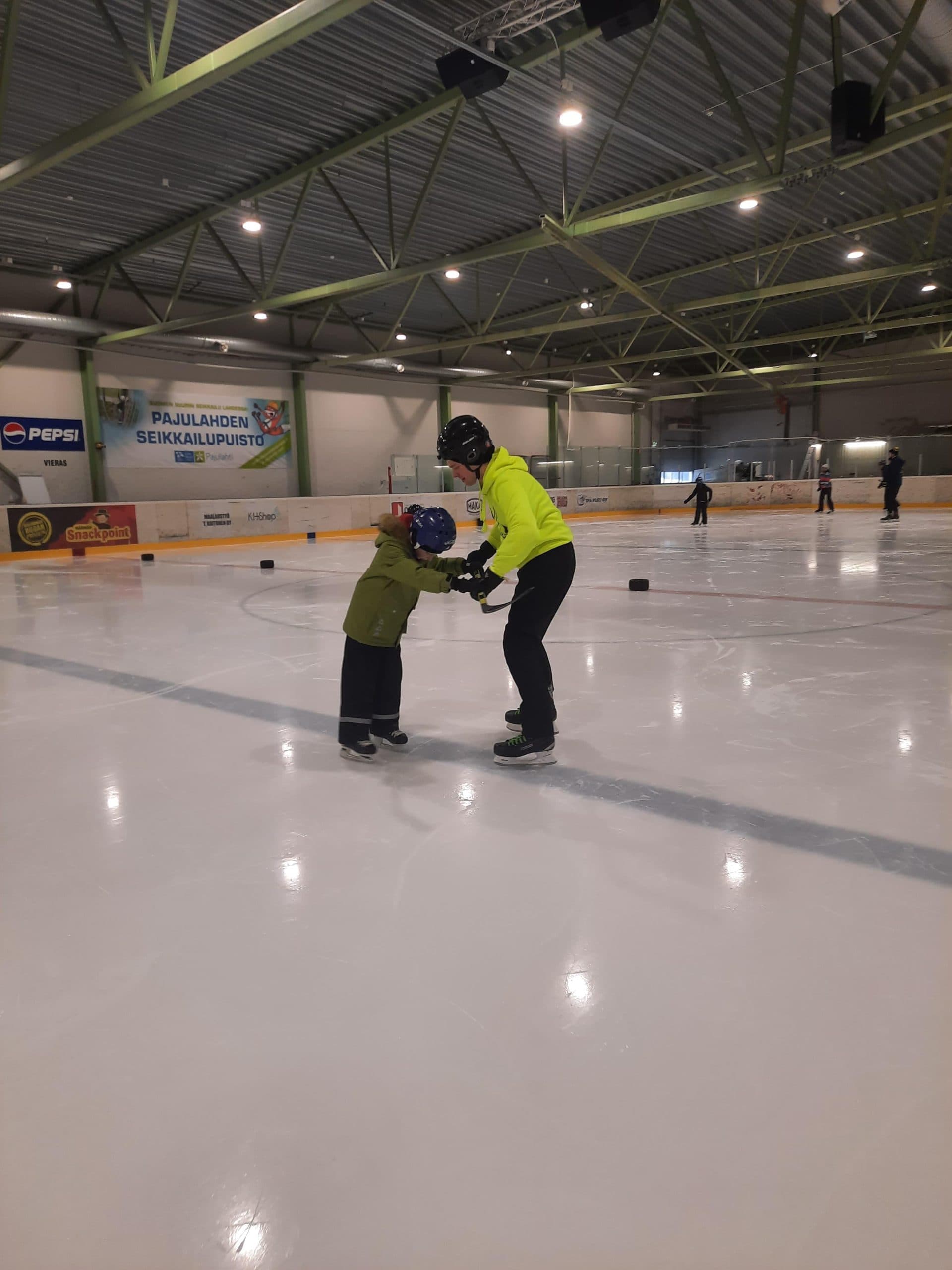 A photo from an ice hockey rink. In the foreground, an instructor in a yellow hoodie holds a stick crossways by the hands, which a child in a green jacket grips while learning to skate. In the background, three young people and one older instructor skate.