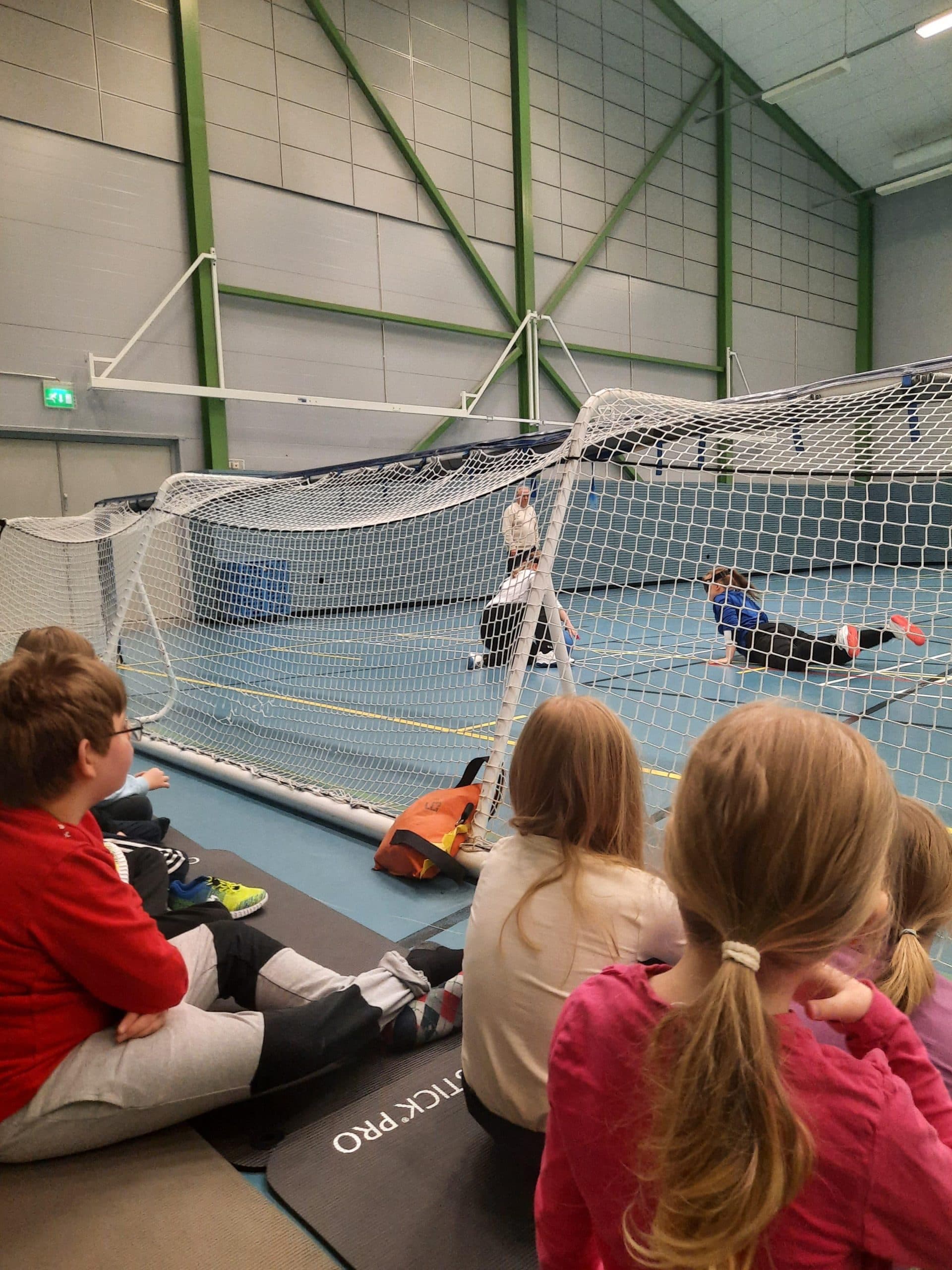 Viisi lasta istumassa maalipallomaalin takana pelihallissa. Kuva otettu lapsien selän takaa. Maalin edessä kaksi pelaajaa torjuvat juuri palloa.