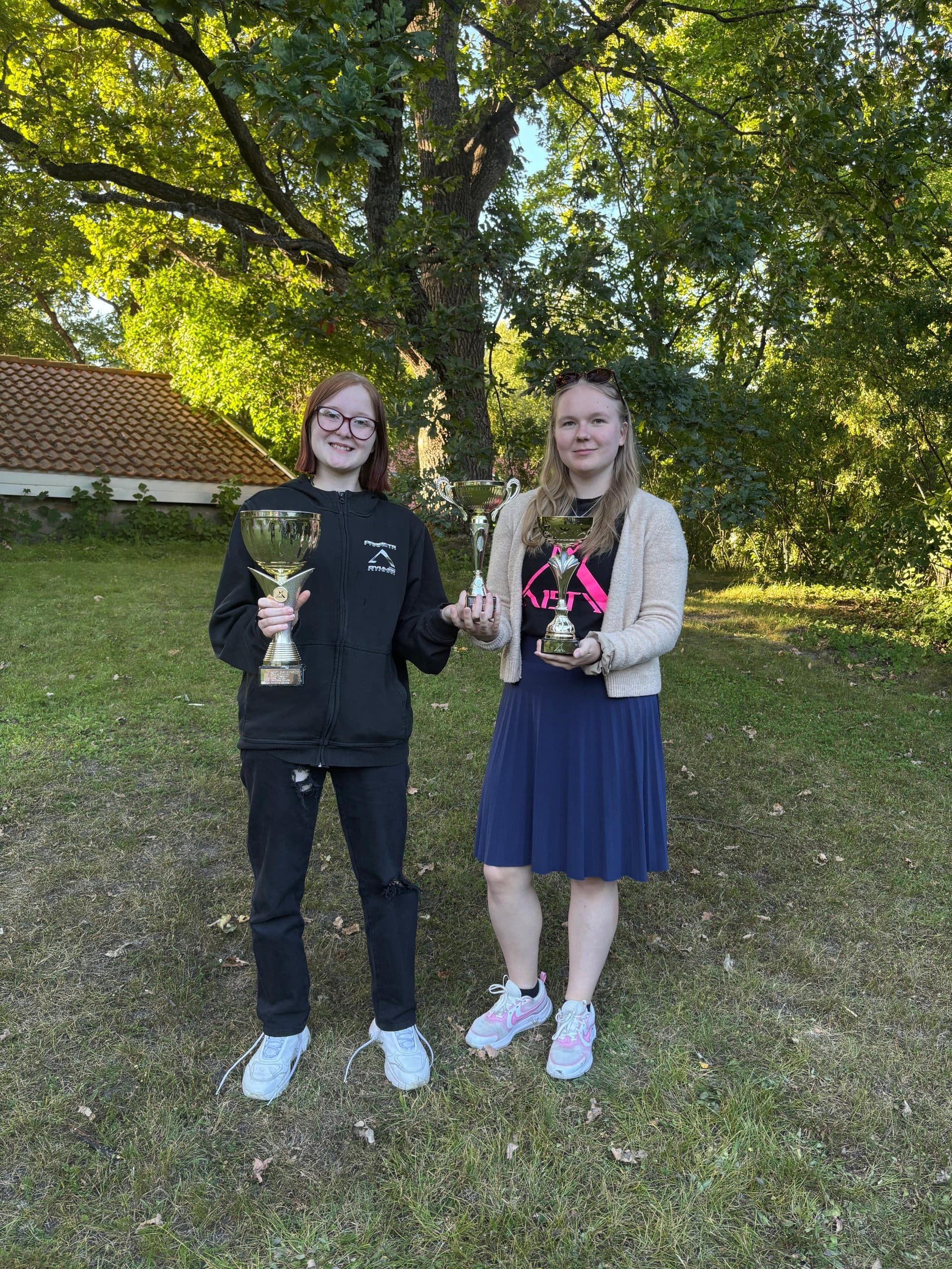 Valo Heinonen mustassa Aistin Uuno juhlahupparissa ja Jasmin Mikkola vaalea neule mustan pinkkilogoisen Aistin t-paidan päällä poseeraamassa nurmikolla. Naisilla on käsissään heidän saaamansa pokaalit ja keskellä he yhdessä pitävät Juuso Somerkiven vuoden vapaaehtois pokaalia.