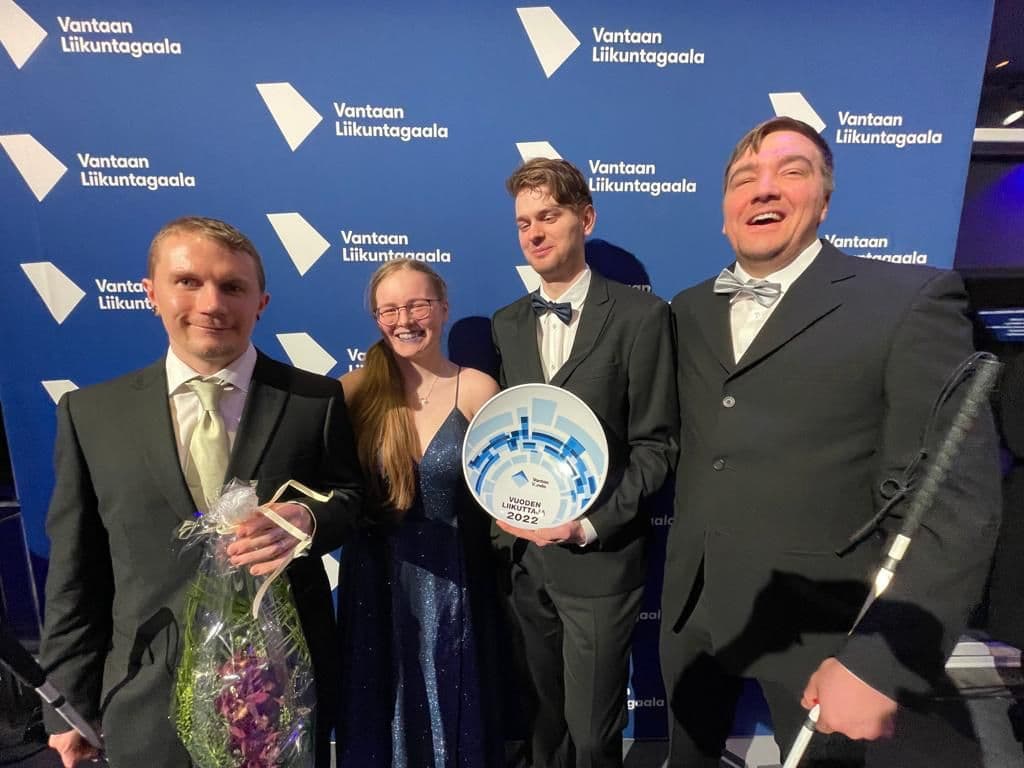 Aistin gaalaedustajat hymyilevät leveästi rinnakkain Vantaan liikuntagaala -pressiseinämän edessä. Miehillä puvut, Henriikalla iltapuku. Krister pitää käsissään palkintolautasta ja Miika kukka-asetelmaa.
