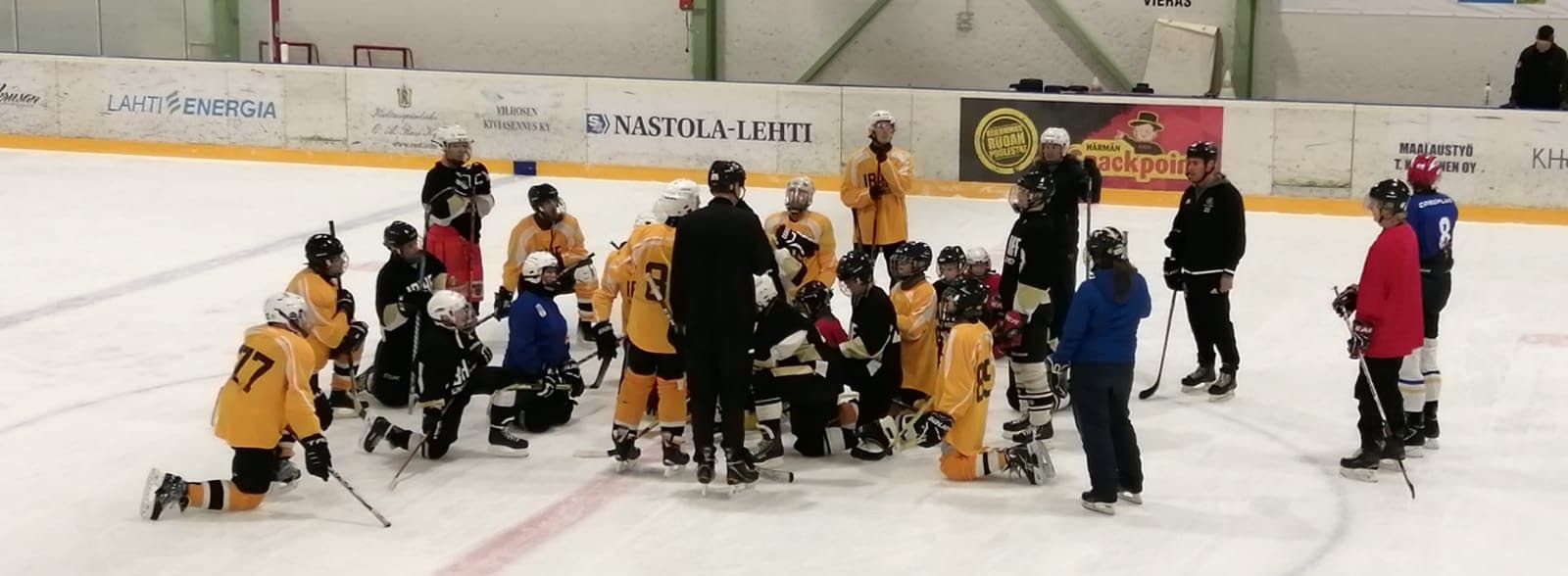A training photo from blind ice hockey where a couple of dozen players stand in the centre circle around the coach, listening to the coach's instructions in the middle.
