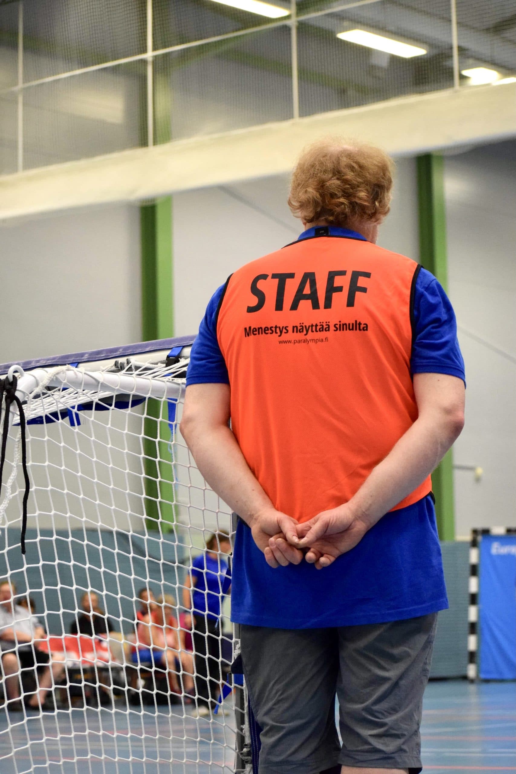 Maalituomari, oranssissa staff-liivissä, selkä kohti kameraa valvomassa maalin vieressä peliä. Sormet pitävät toisistaan kiinni selän takana, jotta olemus olisi ryhdikäs ja siisti. Vieressä maaliverkkoa.