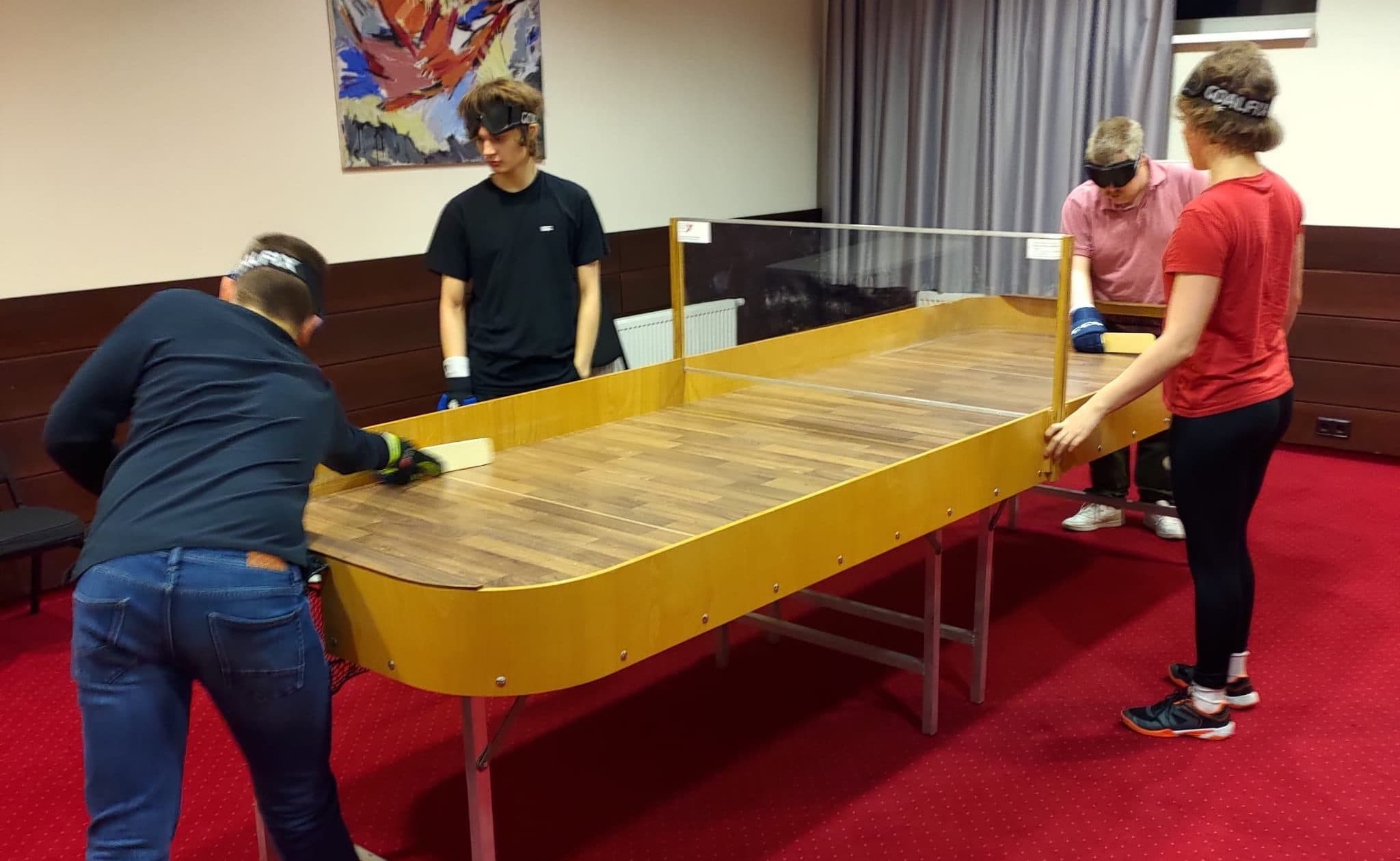 A photo taken from the corner of the table, with two players bent over at either end with wooden bats and black eyeshades, in the flow of the match. The near player is just reaching for the ball with their bat. The table is divided by a transparent plexiglass panel placed across the middle, with a gap underneath for the ball. At the plexiglass, on both sides of the table, two match-watchers have black eyeshades on their foreheads.
