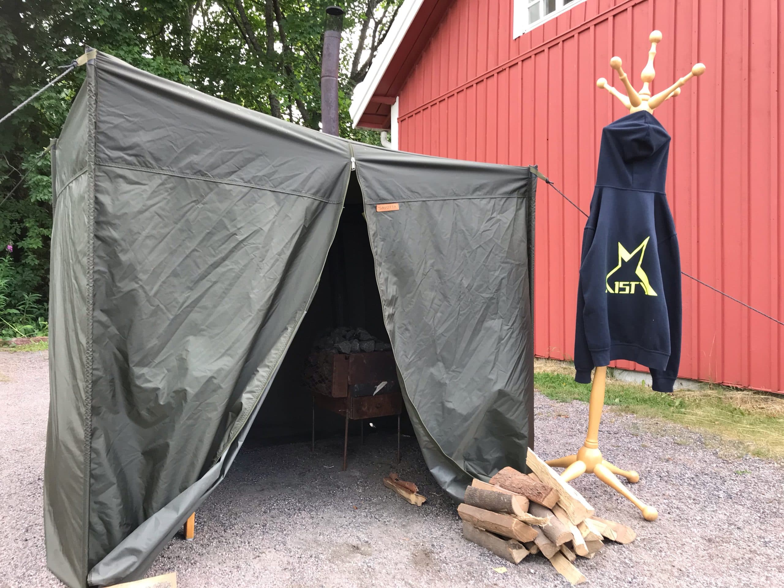 Telttasaunan vetoketjullinen oviaukko aukinaisena. Sisällä näkyy kiuas ja ulkopuolella teltan seinällä roikkuu tumma Aistin huppari isolla Aisti-logolla.